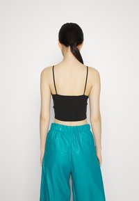 Top cropped preto com alças finas, combinado com calças teal brilhantes de cintura alta. O foco está na vista traseira e no contraste de texturas.