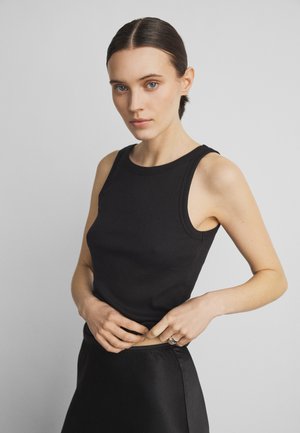 Femeie cu părul legat, purtând un top negru fără mâneci și o fustă, ajustând tivul topului pe un fundal gri simplu.