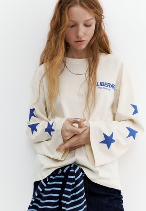 Tânăr cu păr lung roșcat, purtând un hanorac crem cu stele albastre pe mâneci și textul "LIBERATE FREE TO BE YOU," privind în jos.