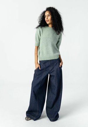 Woman with curly hair wearing a light green short-sleeve sweater and wide-leg dark denim jeans with hands in pockets, looking to the side.