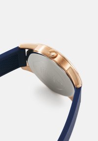 Ceas roz aurit cu carcasă rotundă, având o curea din cauciuc bleu marin și o suprafață texturată. Dispune de o coroană metalică și un capac metalic.