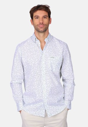 Homme portant une chemise blanche boutonnée avec un petit motif floral bleu et vert, debout les mains dans les poches devant un fond uni.
