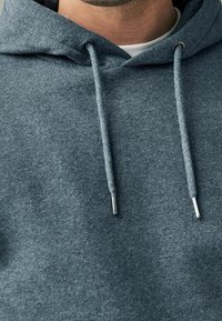 Primo piano di una persona che indossa un cappuccio grigio con cordini e una camicia bianca visibile all'altezza del colletto.