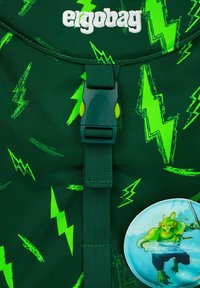 Uno zaino verde presenta un motivo a fulmine, una cinghia anteriore con fibbia e un badge rotondo che mostra un personaggio armato di spada nell'acqua.