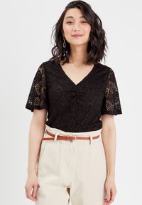 Haut en dentelle noire avec un décolleté en V et des manches courtes, présentant un design froncé au niveau de la poitrine, porté avec un pantalon taille haute beige et une ceinture marron.