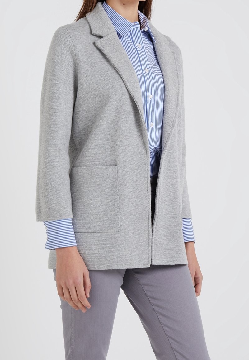 Femme portant un blazer gris clair ouvert sur une chemise rayée bleu et blanc à boutons avec un pantalon gris.