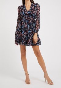 Robe noire à motifs floraux avec manches longues, ourlet volanté et décolleté en V profond. Présente des motifs roses et bleus sur un tissu transparent. Associée à des talons nus.