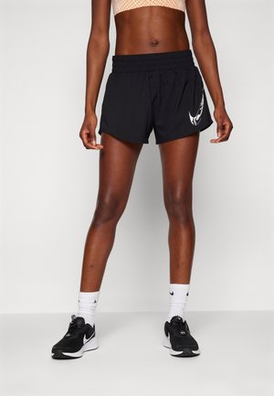 Personne portant un short de sport noir avec un logo blanc, des baskets noires et blanches, des chaussettes blanches et un top court à motifs, debout devant un fond uni.