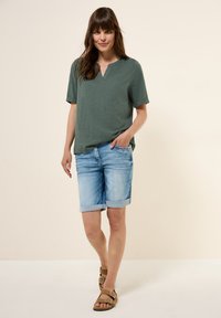 Top verde a maniche corte con scollatura a V, abbinato a pantaloni corti in denim blu chiaro arrotolati ai polsi. Sandali con cinturini e fibbie marroni.
