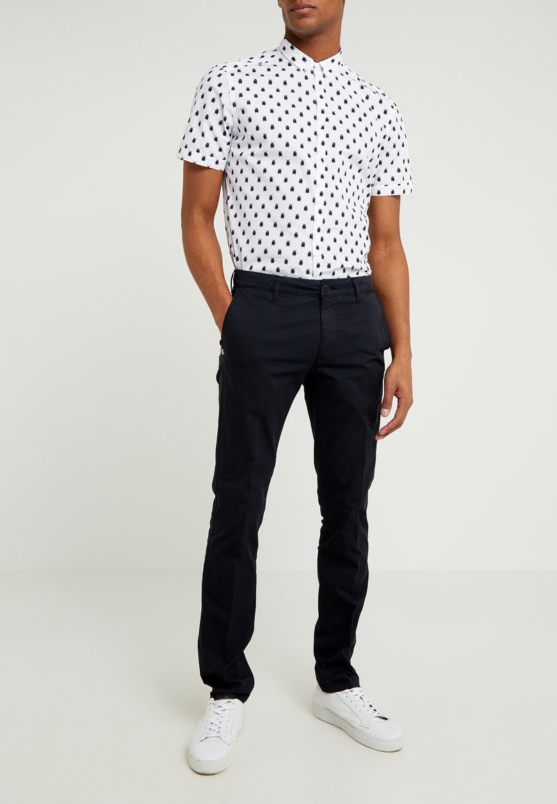 Homme portant une chemise blanche cintrée à manches courtes avec un motif d'abeilles noires, un pantalon noir et des baskets blanches, debout les mains dans les poches.