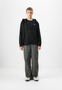 Joven vestido con una sudadera con cremallera negra, pantalones cargo grises y zapatillas blancas, de pie contra un fondo blanco liso, mirando hacia adelante.