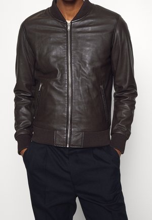 Homme portant une veste bomber en cuir marron foncé avec poignets et taille côtelés, fermeture éclair à l'avant, mains dans les poches.