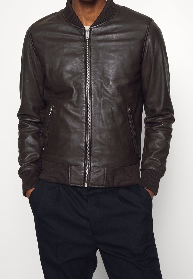 Homme portant une veste bomber en cuir marron foncé avec poignets et taille côtelés, fermeture éclair à l'avant, mains dans les poches.