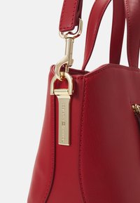 Gros plan d’un sac à main en cuir rouge avec des ferrures métalliques dorées et une étiquette de marque rectangulaire sur la fixation de la bandoulière.