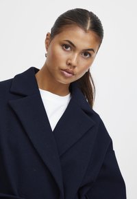 Cappotto in misto lana blu navy con colletto a rever e vestibilità oversize. Presenta una superficie testurizzata ed è indossato su una camicia bianca.