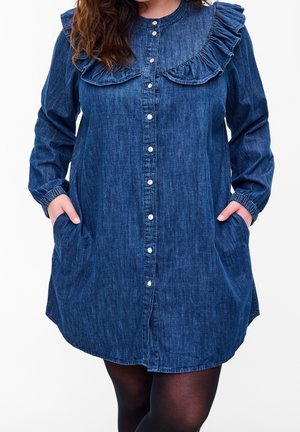 Kobieta ubrana w niebieską sukienkę dżinsową z długimi rękawami, ozdobioną falbanami przy kołnierzu, z przodu zapinaną na guziki, z rękami w kieszeniach, na białym tle.