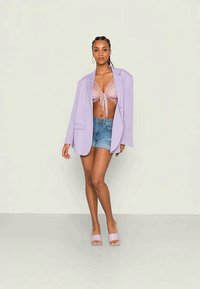 Blazer oversized lilás sobre uma blusa rosa com laço na frente, combinado com calções de ganga e sandálias lilás deslizantes. Texturas suaves e cores suaves.
