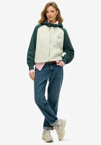 Hooded jack in off-white met donkergroene mouwen, voorzien van een rits, voorkeurszakken en een ontspannen pasvorm, gecombineerd met blauwe spijkerbroeken en sneakers.