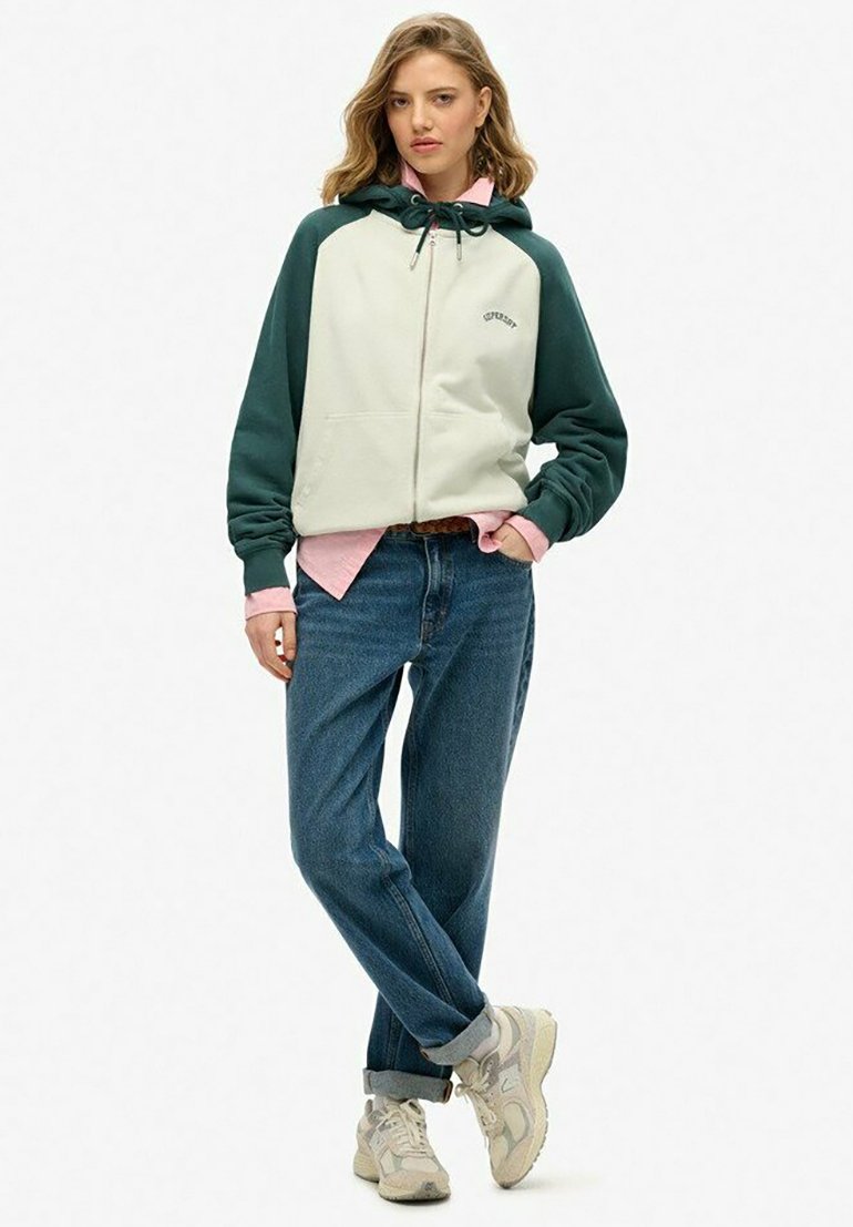 Hooded jack in off-white met donkergroene mouwen, voorzien van een rits, voorkeurszakken en een ontspannen pasvorm, gecombineerd met blauwe spijkerbroeken en sneakers.