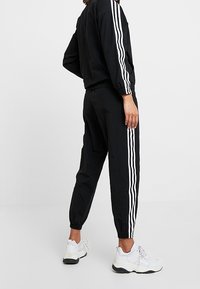 Svart träningsoverall med vita vertikala ränder, med avslappnad passform, elastiska muddar och lätt material. Bärs tillsammans med vita sneakers.