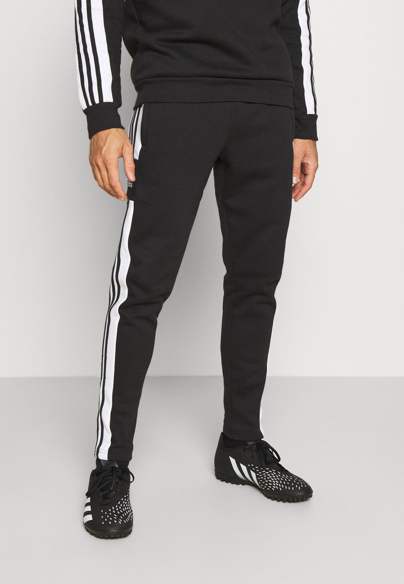 Pantalons de sport noirs avec des rayures blanches sur les côtés, en tissu doux. Associés à des chaussures de sport noires avec un design texturé et des accents blancs.