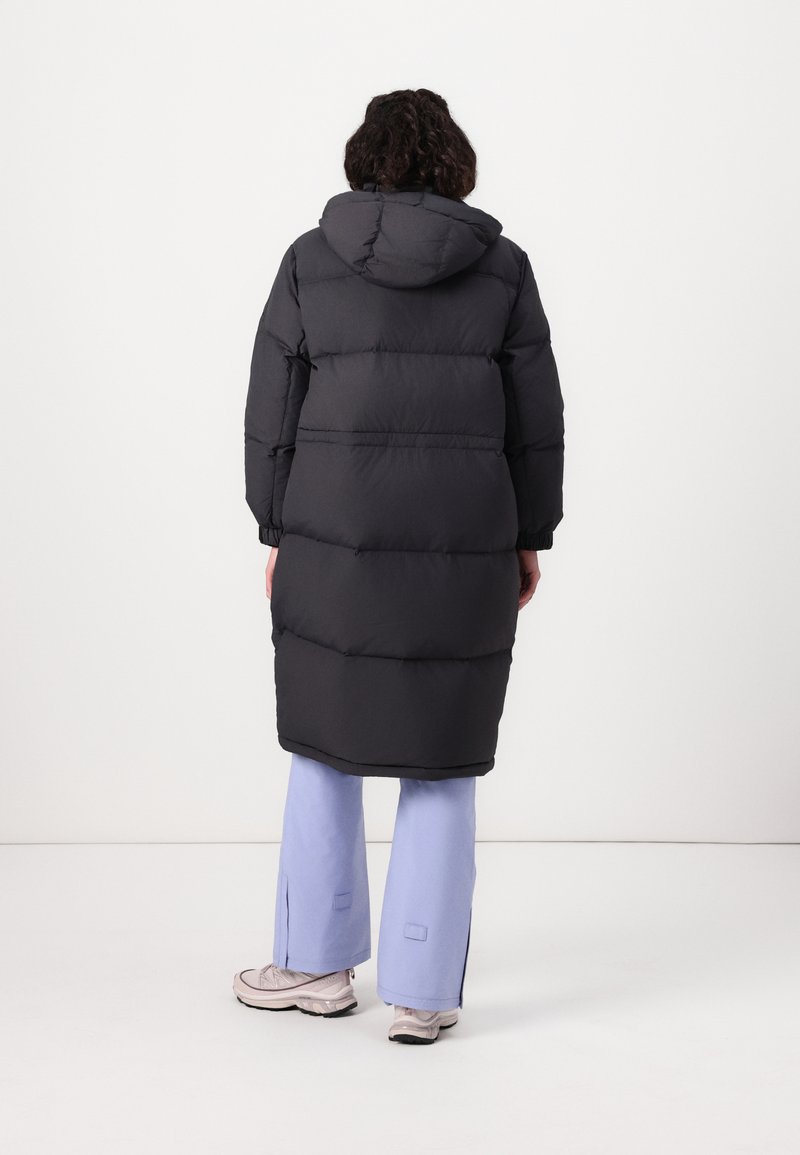 Schwarzer gepolsterter langer Mantel mit Kapuze, ausgestattet mit elastischen Bündchen und tailliertem Schnitt, getragen über hellblauen Hosen und hellen Sneakern.