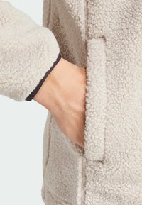 Giacca in pile beige con tessuto strutturato. Dotata di una tasca laterale e di un bordo del polsino nero sulla manica. Design morbido, caldo e casual.