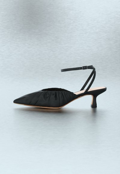 Μαύρο kitten heel με μυτερή μύτη, επάνω μέρος από ρυτιδωμένο ύφασμα και λεπτό λουράκι στον αστράγαλο με πόρπη, εμφανίζεται πάνω σε ανοιχτό γκρι φόντο.