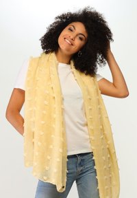 Bufanda de color amarillo claro hecha de un tejido suave y texturizado con acentos anudados. Colgada de manera suelta, presenta un diseño ligero y aireado.