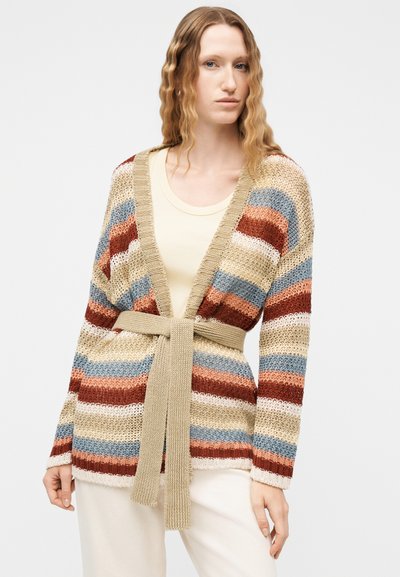 Γυναίκα με κυματιστά μαλλιά φοράει ζακέτα με ζώνη και οριζόντιες ρίγες σε μπεζ, μπλε, σκουριασμένο κόκκινο και λευκό πάνω από μπεζ μπλουζάκι και λευκό παντελόνι.