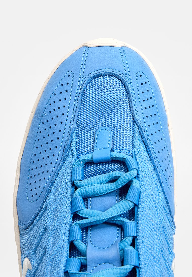 Zapatilla deportiva azul con parte superior de malla, superficie texturizada, perforaciones circulares y cordones planos. Con suela blanca como acento.