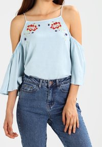 Lyseblå off-shoulder bluse med blomsterbroderi i rød og lilla. Udgiftede ærmer, blødt stof, parret med højtaljede blå jeans.