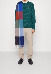 Groene tie-dye sweatshirt, beige broek en zwarte schoenen. Een meerkleurige geruite sjaal hangt omlaag en heeft blauwe, groene en bordeauxrode tinten.