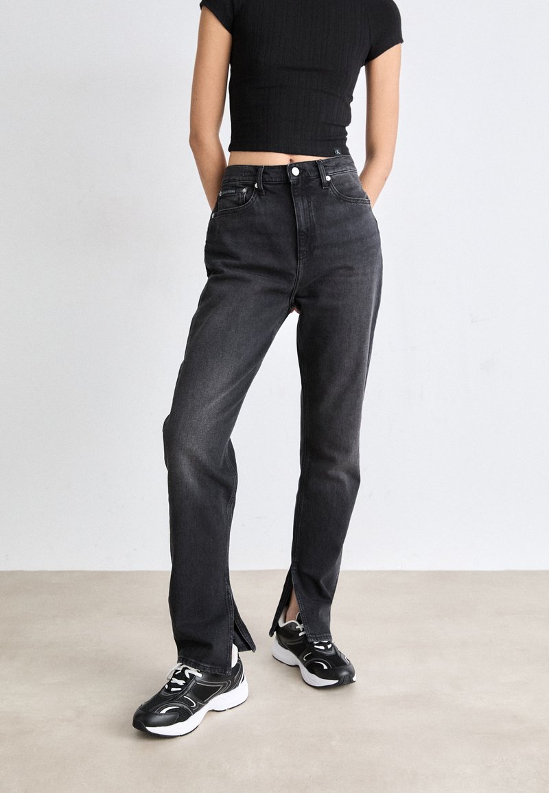 Høytalede svarte denimjeans med sideslisser, kombinert med svart og hvitt sneakers. Stoffet har en glatt tekstur og en klassisk passform.