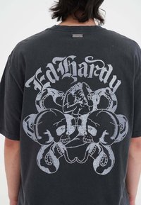 Camiseta de algodón negra con un gran gráfico plateado en la parte posterior, que representa calaveras y una figura entrelazada con serpientes.