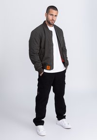 Oliwkowa kurtka typu bomber z prążkowanymi ściągaczami, czerwoną podszewką i naszywką na dole; w połączeniu z czarnymi spodniami cargo i białymi trampkami.