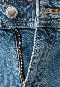 Jean en denim bleu partiellement dézippé montrant un bouton en métal et une fermeture éclair visible avec des surpiqûres orange autour de la taille.
