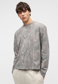 Noor mees tumedate juustega, kellel on seljas halli mustriga pika varrukaga särk ja heledad beežid püksid, seisab valgel taustal käed taskutes.