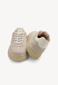 Sneakers in suede beige con accenti bianchi, punta rotonda, lacci piatti e suole spesse e testurizzate. Presentano un retro imbottito e un design sobrio.