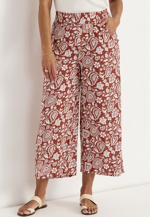 Kobieta ubrana w szerokie spodnie w kolorze rdzy z białym kwiatowym wzorem paisley, elastycznym pasem, bocznymi kieszeniami i kremowymi sandałami.
