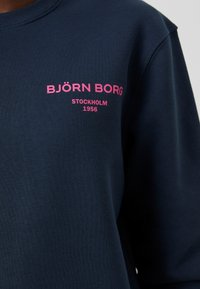Námořnická modrá mikina vyrobená z bavlny, s růžovým textem "BJÖRN BORG STOCKHOLM 1956" na levé části hrudi. Jednoduchý design, kulatý výstřih.