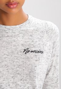 Hellgrauer Pullover mit einem strukturierten Muster, auf dessen Vorderseite schwarzer gestickter Text mit der Aufschrift "No worries" prangt. Rundhalsausschnitt und lässiger Schnitt.