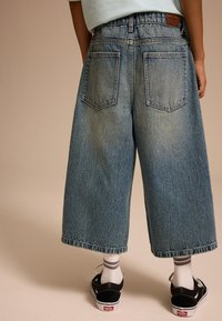 Culottes en denim bleu à coupe ample, avec deux poches arrière et une coupe droite. Portées avec des chaussures noires et des chaussettes blanches avec des rayures grises.