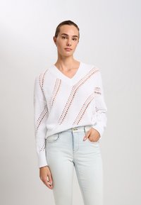 Weißer, gestrickter Pullover mit V-Ausschnitt, der ein strukturiertes Muster aus großen und kleinen Ausschnitten aufweist. Kombiniert mit hellblauen, hochtaillierten Jeans.
