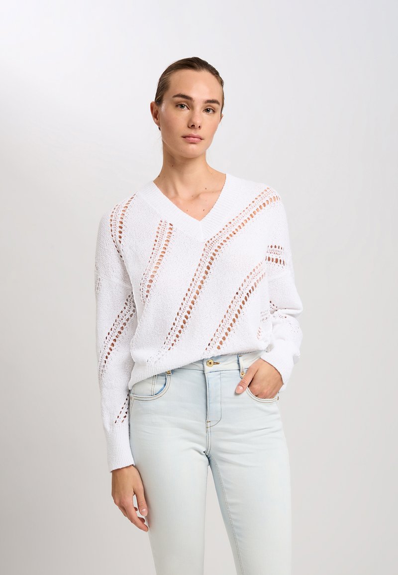 Weißer, gestrickter Pullover mit V-Ausschnitt, der ein strukturiertes Muster aus großen und kleinen Ausschnitten aufweist. Kombiniert mit hellblauen, hochtaillierten Jeans.