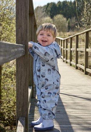 Bambino piccolo con tuta impermeabile azzurro chiaro a stampa pirata e stivali bianchi che sorride, appoggiato alla ringhiera di un ponte pedonale di legno in un ambiente esterno soleggiato.
