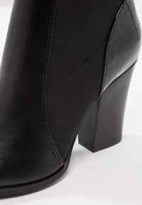 Bottine en cuir noir et tissu avec un talon bloc, coutures lisses et bout arrondi, présentée sur un fond blanc.