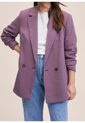 Femme portant un blazer pied-de-poule violet et noir sur une chemise blanche avec un jean taille haute bleu, debout devant un fond beige.