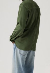 Camicia a maniche lunghe verde oliva con vestibilità comoda, caratterizzata da un orlo arrotondato e polsini con bottoni, abbinata a pantaloni in denim blu chiaro.