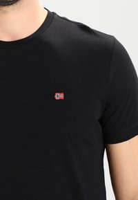 T-shirt nera in cotone a maniche corte, con un piccolo logo della bandiera ricamato in rosso e bianco sul petto. Design classico con collo a giro.